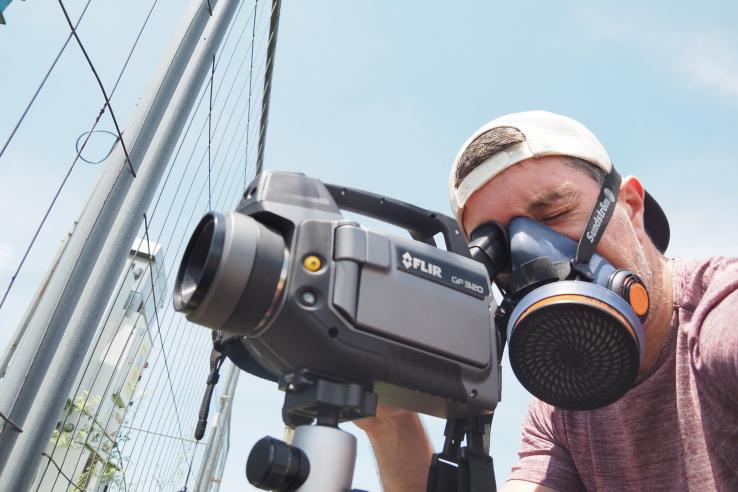 James, wearing a gas mask, looks at an oil well through a camera