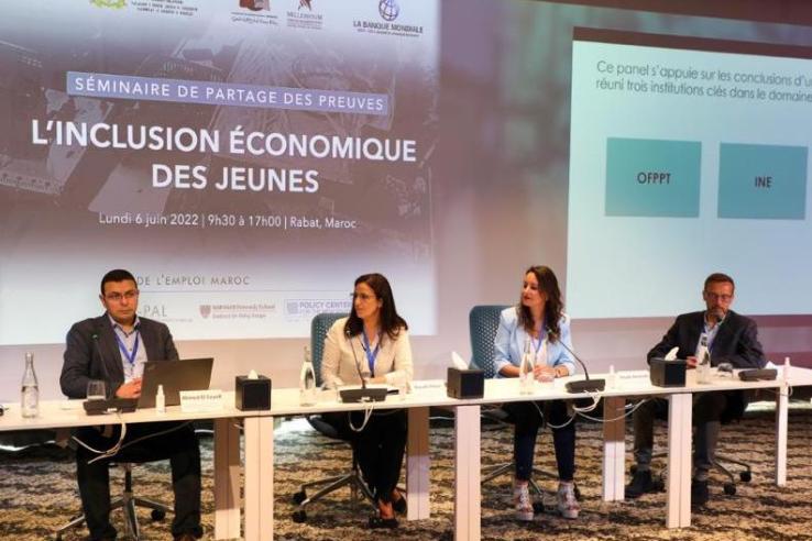 Four speakers are seated at a panel table at the front of a room.