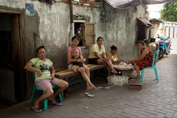Three women are seated on a bench outside of a dwelling, one of whom holds a small child. Two women are seated next to the bench, peeling garlic over a large bowl.