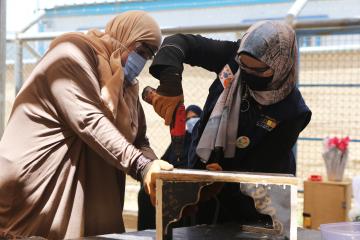 Two women wearing headscarves use a drill to upcycle a piece of wood furniture.